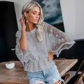 A blonde model showcasing a semi-sheer light gray lace top featuring dramatic batwing sleeves and intricate floral embroidery, styled with casual denim shorts in an indoor setting